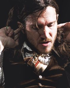 a man with long hair wearing a vest and bow tie holding his hands to his ears