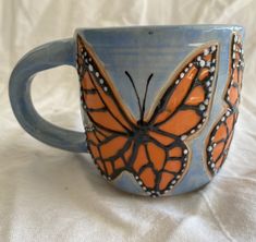 a blue and orange coffee cup with a butterfly on it's side sitting on a white sheet