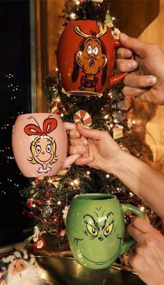 two people holding up mugs with cartoon faces on them next to a christmas tree