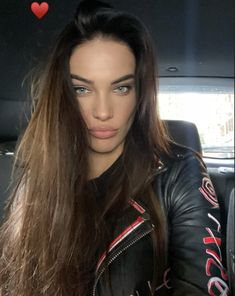 a woman with long brown hair sitting in the back seat of a car wearing a leather jacket