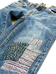 a pair of blue jeans with an embroidered tree on the front and back pocket, sitting on a white surface