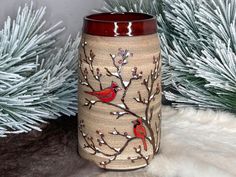 a vase with two red birds on it sitting in front of some pine needles and branches