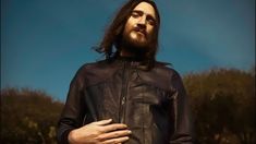 a man with long hair and beard wearing a black leather jacket standing in a field