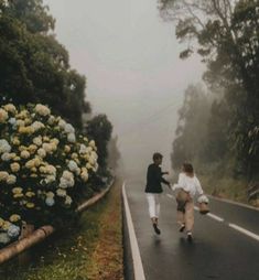 two people are walking down the road in the middle of the day with flowers on either side