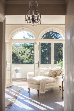 the sun shines through two large windows into a living room with white furniture and chandelier