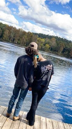 a man and woman standing on a dock looking at the water with trees in the background