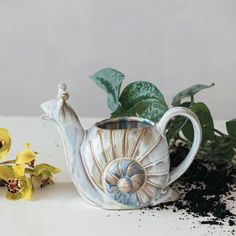 a ceramic teapot with a snail on it next to some flowers and dirt in the background