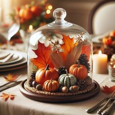 a glass clochel with fall leaves and pumpkins under it on a table