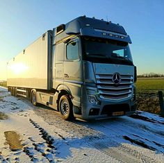 a semi truck driving down a snow covered road next to a green grass and tree