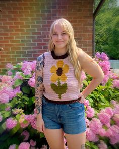 a woman standing in front of pink flowers wearing a crocheted top with sunflowers on it