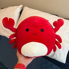 a hand holding a red and white stuffed animal on top of a bed next to pillows