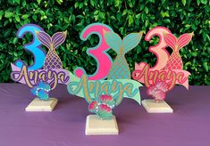 three wooden numbers with mermaid tails and flowers on top of a purple tablecloth covered table