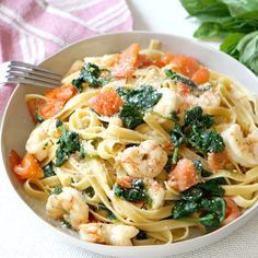 a white bowl filled with pasta and shrimp on top of a table next to spinach leaves