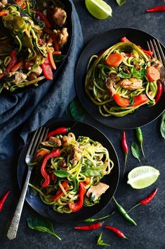 two black plates filled with noodles and vegetables on top of a blue cloth next to lime wedges