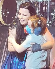 a woman holding a child in her arms on stage with drums behind her and another person standing next to her