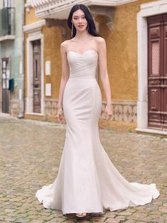 a woman in a white wedding dress standing on a cobblestone street with buildings behind her