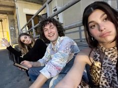 three young people sitting on the ground in front of a building and one is holding a cell phone