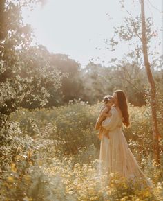 a woman holding a small dog in a field with tall grass and trees behind her