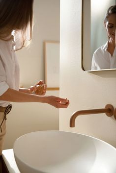 a woman is holding something in her hand while standing next to a sink and mirror