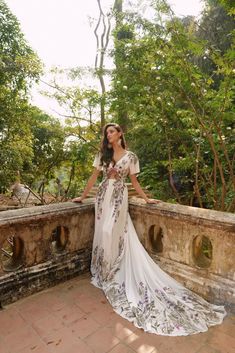 a woman in a long dress standing on a stone wall with trees and bushes behind her