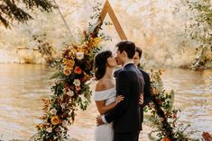 a bride and groom kissing in front of an arch decorated with flowers on the riverbank