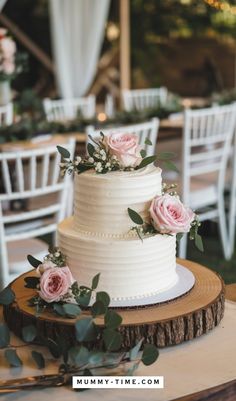 a white wedding cake with pink roses and greenery sits on a wooden slice in front of chairs