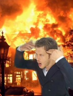 a man holding his head in front of a large fire that is burning behind him