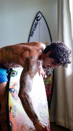 a man standing next to a surfboard with tattoos on his arm and chest,