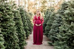 a pregnant woman in a long red dress standing between rows of christmas tree's