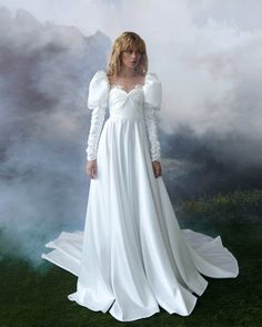 a woman in a white dress standing on top of a grass covered field with clouds behind her