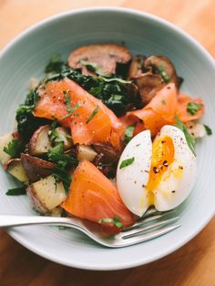 a bowl filled with vegetables and eggs on top of a wooden table next to a fork