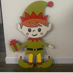 a wooden cutout of a elf holding a present box in one hand and standing next to a wall