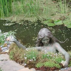 a statue of a mermaid holding a fish in a pond surrounded by grass and flowers