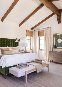 a bedroom with white bedding, green headboard and footstool in front of windows