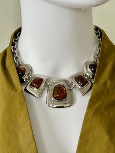 This Vintage Kiam Family Faux Amber Necklace was sourced in Los Angeles!  The modernist silvertone statement necklace is made up of an anchor chain on either side, and enclosed with a lobster claw clasp. In the center, there are graduating highly polished silvertoned linked plates (in various rounded polygon shapes) with central smooth cabochon stones made to look like faux amber. The "stones" are quite luminous and the colors shifts from a lovely orange to a deep brown.  A "Kiam Family" hang tag hangs from one end.  Elena Kiam is the creative director behind "Lia Sophia" which started after Kiam family jewelry in the 1970's. Lia Sophia jewelry was started in 1986. The company sold its jewelry mostly at home parties so it is considered rare and hard to come by!   -Total length: 16 in (but Polygon Shapes, Family Jewelry, Home Parties, Anchor Chain, Family Jewellery, Lia Sophia, Bib Necklaces, Amber Necklace, Amber Stone