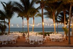 tables and chairs are set up under palm trees on the beach at dusk with string lights strung over them