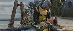 two men in yellow and black diving suits are on the beach while one man is laying down