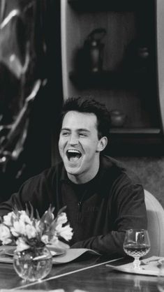 a man laughing while sitting at a table in front of a vase with flowers on it