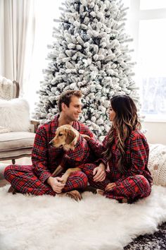 a man and woman in matching pajamas sitting next to a christmas tree with their dog