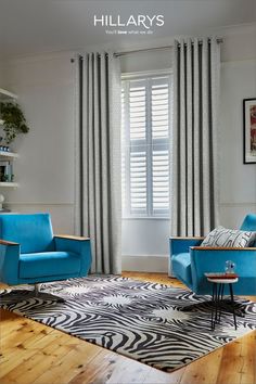 Two bright blue armchairs that frame a window that is dressed with shutters and curtains. The curtains are floor to ceiling with a geometric pattern. There is a zebra print rug on the floor and a small coffee table next to one of the chairs. Curtains With Shutters, Shutters Bay Window, Shutters With Curtains, Shutter Styles, Bay Window Shutters, Cafe Style Shutters, False Wall, Natural Stain Wood, Shaped Windows