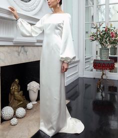 a woman standing in front of a fire place wearing a white dress with long sleeves