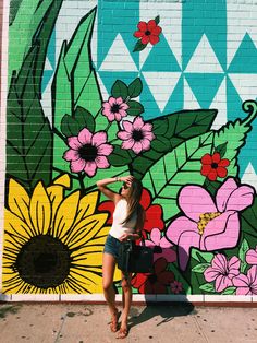 a woman standing in front of a colorful wall with flowers painted on it and holding a black purse