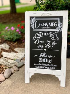 a chalkboard sign sitting on the side of a sidewalk next to flowers and trees