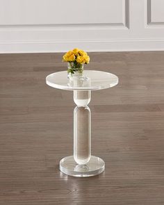 a clear glass table with yellow flowers in the center on a wooden floor next to a white door