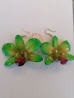 two green orchids are hanging from silver earwires on a white surface with a monogram in the background