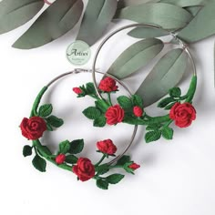 a pair of hoop earrings with red roses on them and green leaves around the hoop