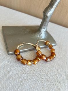 These stunning warm bronze copper tone hoop earrings feature beautiful shiny acrylic beads with a pearlescent finish, creating a unique metallic glow.  The rich brown hues combined with a touch of gold add an elegant, earthy charm to these statement hoops. Perfect for adding a sophisticated flair to any outfit. Ideal for those who love bold yet versatile accessories, these brown pearlescent hoops make a thoughtful gift for her, whether for a special occasion or simply as a stylish addition to her jewelry collection. Length: 1 3/4 inches  Width: 2 inches Thoughtful Gifts For Her, Hoops Gold, Bronze Earrings, Touch Of Gold, Copper Earrings, Jewelry Earrings Hoops, Acrylic Beads, Jewelry Collection, Jewelry Earrings