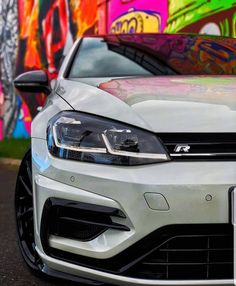 the front end of a white car parked in front of graffiti covered wall with colorful spray paint on it