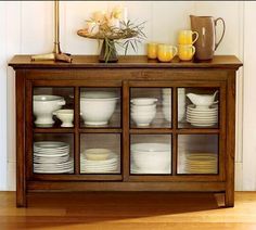 a wooden cabinet filled with white plates and cups next to a vase on top of a table