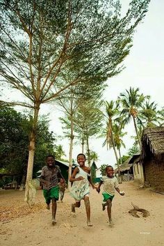 three young children running in the dirt near some trees and shacks with thatched roof